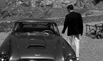 Movie still from “Phaedra” (1962), directed by Jules Dassin – A black and white photo of a man standing next to a car; Wide shot, High angle