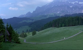 Movie still from “Phenomena” (1985), directed by Dario Argento – A road going through a lush green field with mountains in the background; Extreme Wide shot, High angle