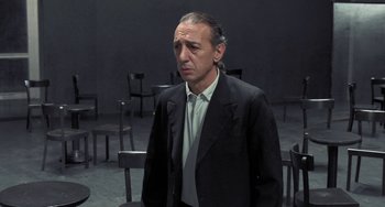 Movie still from “Pina” (2011), directed by Wim Wenders – A man wearing a suit and tie standing in front of some chairs; Medium shot, Over the shoulder angle
