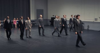 Movie still from “Pina” (2011), directed by Wim Wenders – A group of men in business suits walking across a stage; Wide shot, High angle