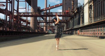 Movie still from “Pina” (2011), directed by Wim Wenders – A woman in a black and white dress dancing in a parking lot; Wide shot, Low angle