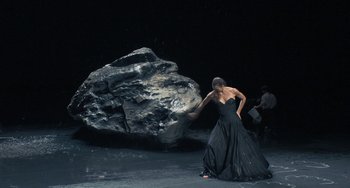 Movie still from “Pina” (2011), directed by Wim Wenders – A woman in a black dress is standing next to a large rock; Wide shot, Low angle