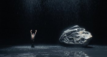 A person standing in the middle of the water in front of a large rock