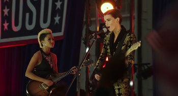 Movie still from “Pitch Perfect 3” (2017), directed by Trish Sie – A woman singing and playing a guitar in front of another woman; Medium shot, Over the shoulder angle