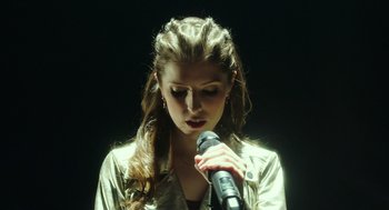 Movie still from “Pitch Perfect 3” (2017), directed by Trish Sie – A woman holding a microphone in front of a black background; Close Up shot, Overhead angle