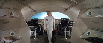 Movie still from “Planet of the Apes” (1968), directed by Franklin J. Schaffner – A man in a white suit standing in front of chairs in a room; Wide shot, High angle