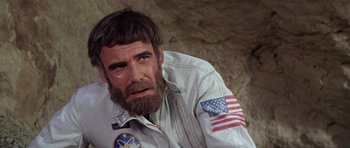 Movie still from “Planet of the Apes” (1968), directed by Franklin J. Schaffner – A man with a beard wearing a white jacket with an american flag on it; Close Up shot, Over the shoulder angle
