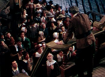 Movie still from “Plymouth Adventure” (1952), directed by Clarence Brown – A group of people sitting in a room with a man standing on a boat; Wide shot, High angle