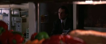 Movie still from “Point of No Return” (1993), directed by John Badham – A man in a suit and tie holding a camera in front of vegetables; Close Up shot, Low angle
