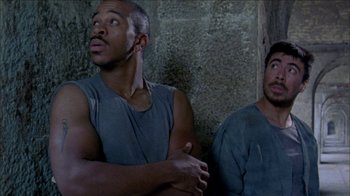Movie still from “Poison” (1991), directed by Todd Haynes – Two men sitting next to each other in front of a stone wall; Medium shot, Low angle