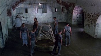 Movie still from “Poison” (1991), directed by Todd Haynes – A group of men standing in a room next to an old structure; Wide shot, High angle