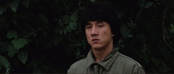 Movie still from “Police Story” (1985), directed by Jackie Chan – A young man with a black wig is standing in front of a tree; Close Up shot, Low angle