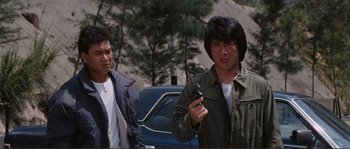 Movie still from “Police Story” (1985), directed by Jackie Chan – A man holding a gun while standing next to another man in front of a car; Medium shot, Low angle