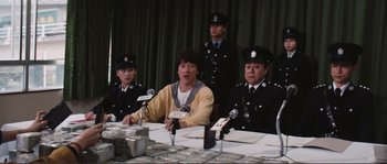 Movie still from “Police Story” (1985), directed by Jackie Chan – A group of men sitting at a table with microphones; Medium shot, Low angle