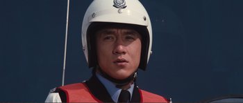 Movie still from “Police Story” (1985), directed by Jackie Chan – A police officer wearing a white helmet and a red vest; Close Up shot, Low angle