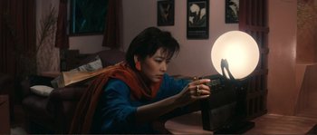Movie still from “Police Story” (1985), directed by Jackie Chan – A woman sitting at a table looking at an object in front of a lamp; Medium shot, High angle