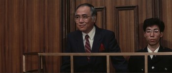 Movie still from “Police Story” (1985), directed by Jackie Chan – A man in a suit and tie sitting in front of a wooden wall; Medium shot, High angle