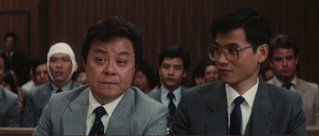 Movie still from “Police Story” (1985), directed by Jackie Chan – A group of men in suits sitting in front of a wall; Close Up shot, Over the shoulder angle