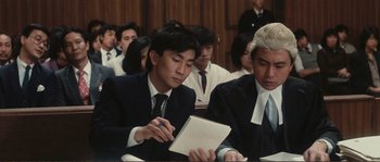 Movie still from “Police Story” (1985), directed by Jackie Chan – A man in a suit and tie writing on a notepad in front of other men; Medium shot, High angle
