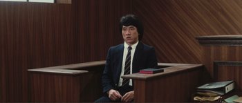 Movie still from “Police Story” (1985), directed by Jackie Chan – A man in a suit and tie sitting in a court room; Medium shot, High angle