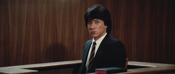 Movie still from “Police Story” (1985), directed by Jackie Chan – A man in a suit and tie sitting in front of a book; Medium shot, Low angle