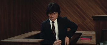 Movie still from “Police Story” (1985), directed by Jackie Chan – A man in a suit and tie sitting in a court room; Medium shot, Low angle