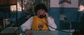 Movie still from “Police Story” (1985), directed by Jackie Chan – A man sitting at a table talking on a phone; Medium shot, High angle