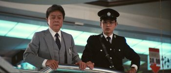 Movie still from “Police Story” (1985), directed by Jackie Chan – A man and a man in a suit standing next to each other in front of an escalator; Medium shot, Low angle
