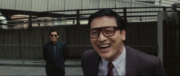 Movie still from “Police Story 2” (1988), directed by Jackie Chan – A man wearing glasses and a suit and tie smiling; Close Up shot, Over the shoulder angle