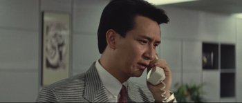 Movie still from “Police Story 2” (1988), directed by Jackie Chan – A man talking on a phone while wearing a suit and tie; Close Up shot, Low angle