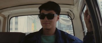 Movie still from “Police Story 2” (1988), directed by Jackie Chan – A young man wearing sunglasses in the back of a car; Close Up shot, Over the shoulder angle