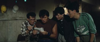 Movie still from “Police Story 2” (1988), directed by Jackie Chan – A group of men standing around looking at a piece of paper; Medium shot, High angle