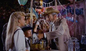 Movie still from “Pollyanna” (1960), directed by David Swift – An old man in a suit and a cowboy hat is serving a young boy at an amusement park; Medium shot, Over the shoulder angle
