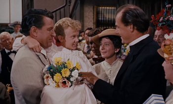 Movie still from “Pollyanna” (1960), directed by David Swift – A group of people gathered around a little girl in a white dress; Medium shot, Over the shoulder angle