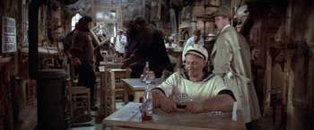 Movie still from “Popeye” (1980), directed by Robert Altman – A man sitting at a table with a glass of beer; Wide shot, High angle