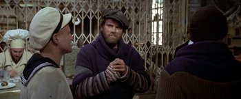 Movie still from “Popeye” (1980), directed by Robert Altman – A man in a hat and sweater clasps his hands in prayer; Medium shot, Over the shoulder angle