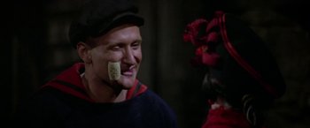 Movie still from “Popeye” (1980), directed by Robert Altman – A man wearing a black hat with a piece of food in his mouth; Close Up shot, Over the shoulder angle