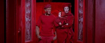 Movie still from “Popeye” (1980), directed by Robert Altman – A man and woman holding a baby in a red outfit; Medium shot, High angle