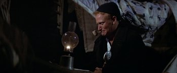 Movie still from “Popeye” (1980), directed by Robert Altman – A man smoking a pipe next to an oil lamp; Medium shot, Low angle