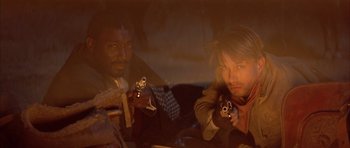 Movie still from “Posse” (1993), directed by Mario Van Peebles – Two men are holding guns in a dimly - lit room; Close Up shot, High angle
