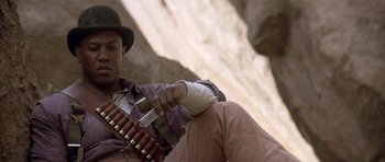 Movie still from “Posse” (1993), directed by Mario Van Peebles – A man sitting on the ground holding a rifle; Close Up shot, Low angle