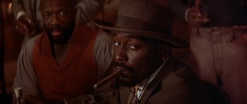 Movie still from “Posse” (1993), directed by Mario Van Peebles – A man in a hat and a suit smoking a cigar; Close Up shot, Low angle