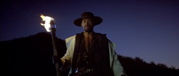 Movie still from “Posse” (1993), directed by Mario Van Peebles – A man in a hat holding a stick in the dark; Medium shot, Low angle