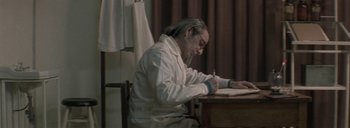 Movie still from “Post Mortem” (2010), directed by Pablo Larraín – An older man sitting at a desk writing on a piece of paper; Medium shot, Low angle