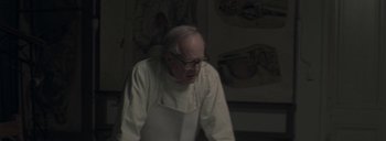 Movie still from “Post Mortem” (2010), directed by Pablo Larraín – An older man wearing a white apron and glasses; Medium shot, Over the shoulder angle