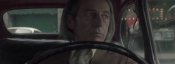 Movie still from “Post Mortem” (2010), directed by Pablo Larraín – A man in a suit and tie sitting in the driver's seat of a car; Close Up shot, Low angle