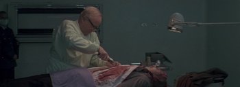 Movie still from “Post Mortem” (2010), directed by Pablo Larraín – An old man is cutting a woman's arm; Medium shot, High angle