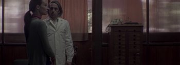 Movie still from “Post Mortem” (2010), directed by Pablo Larraín – A woman in a white lab coat standing in front of a desk; Medium shot, Low angle