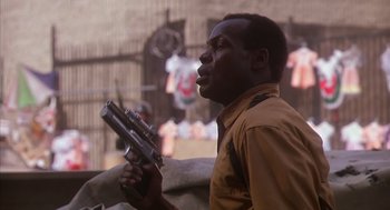 Movie still from “Predator 2” (1990), directed by Stephen Hopkins – A man holding a gun while standing next to a couch; Close Up shot, Low angle