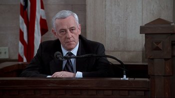 Movie still from “Primal Fear” (1996), directed by Gregory Hoblit – An older man sitting at a podium in front of a microphone; Medium shot, Low angle